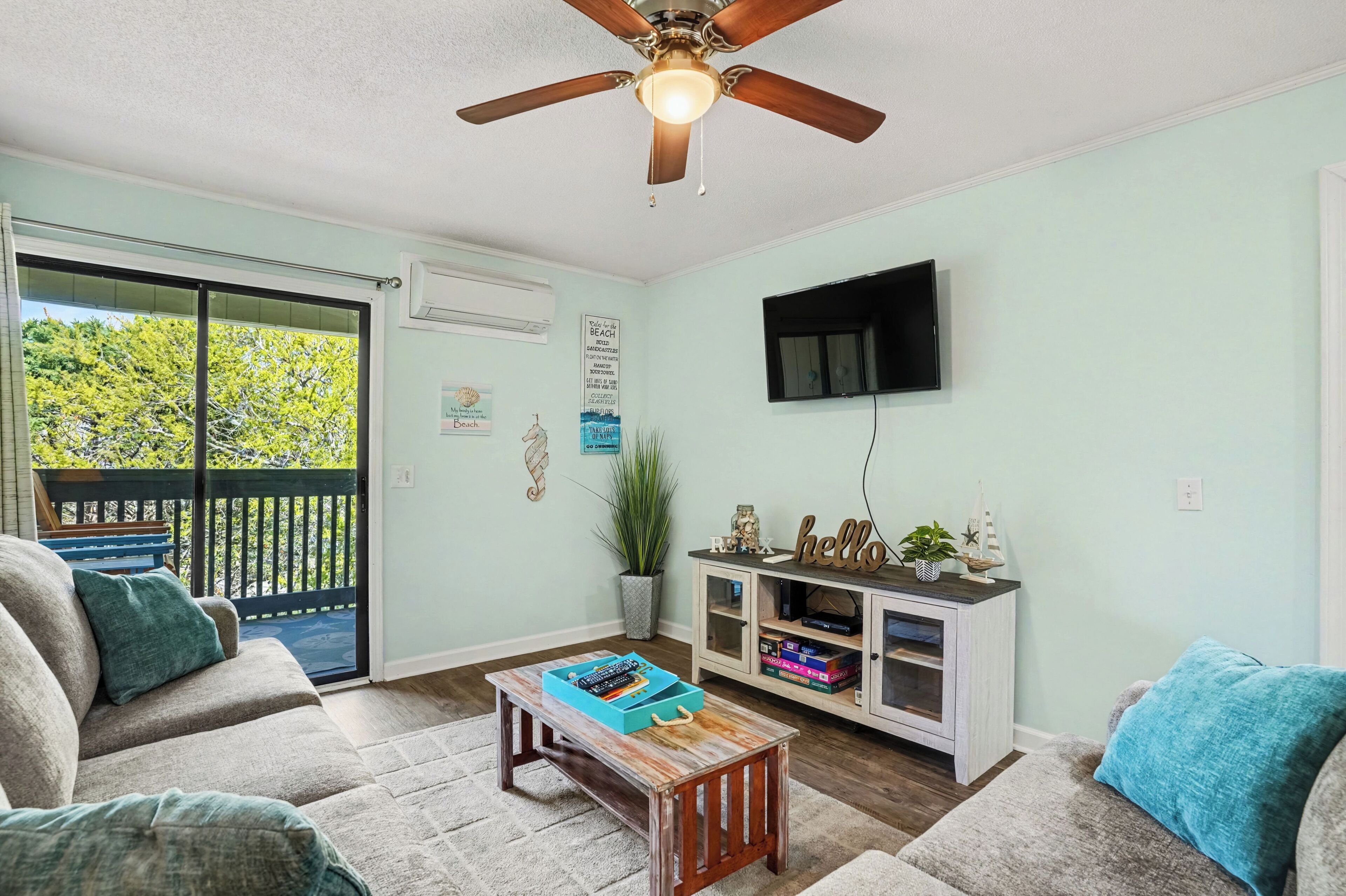 A living room with a ceiling fan, a TV mounted on the wall, a sliding glass door to the outside, a small wooden coffee table, and a white entertainment unit with decorative items.