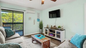 A living room with a ceiling fan, a TV mounted on the wall, a sliding glass door to the outside, a small wooden coffee table, and a white entertainment unit with decorative items. - A Place At The Beach #3C Short Walk to the Beach! (North Myrtle Beach)