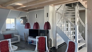 Un salon moderne avec un canapé blanc, des fauteuils rouges, une table basse en verre et un téléviseur. Un escalier avec des rampes blanches mène à un niveau supérieur.