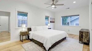 Un dormitorio con una cama grande, dos mesitas de noche con lámparas, un ventilador de techo y ventanas amplias con cortinas.