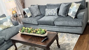 A living room with a grey sofa, a wooden coffee table with a plant, and a remote control on it. - Nice family home (Hiram)