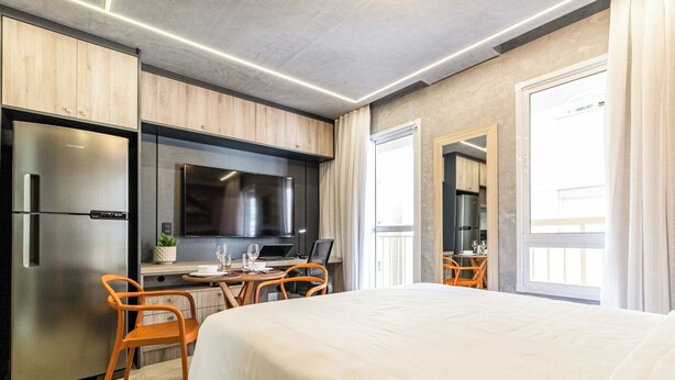 Una habitación de hotel moderna con una cama grande, una kitchenette con refrigerador y estufa, un comedor con sillas y un ventanal amplio.