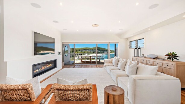 Un salón moderno con un sofá grande, una chimenea y vistas a una playa y colinas a través de amplios ventanales.