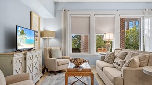 Un salon avec un canapé beige, une table basse en verre, un téléviseur posé sur un meuble et une vue sur les arbres à travers de grandes fenêtres.
