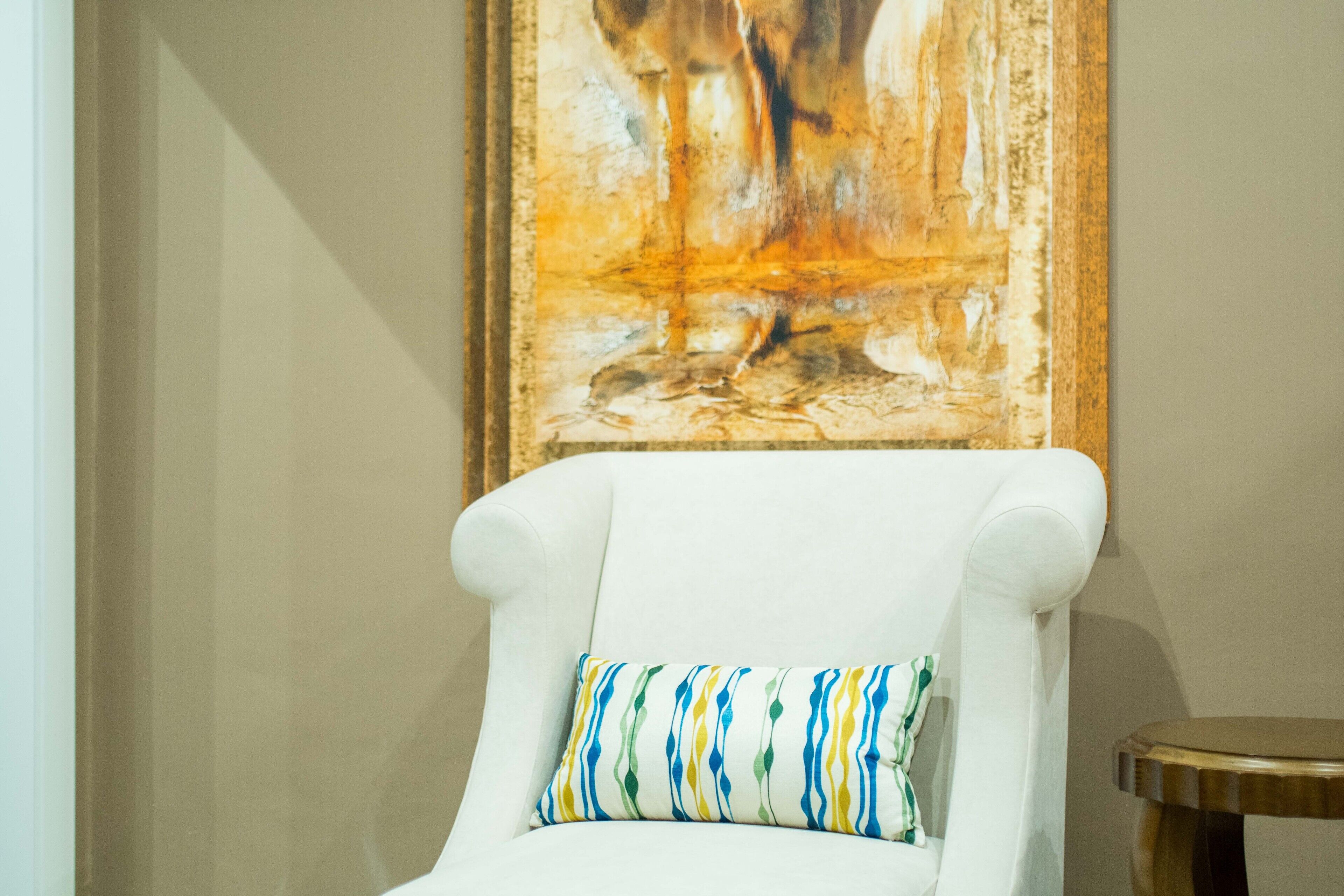 Een witte fauteuil met een kleurrijk kussen, een rond houten krukje, en een groot abstract schilderij aan de muur.