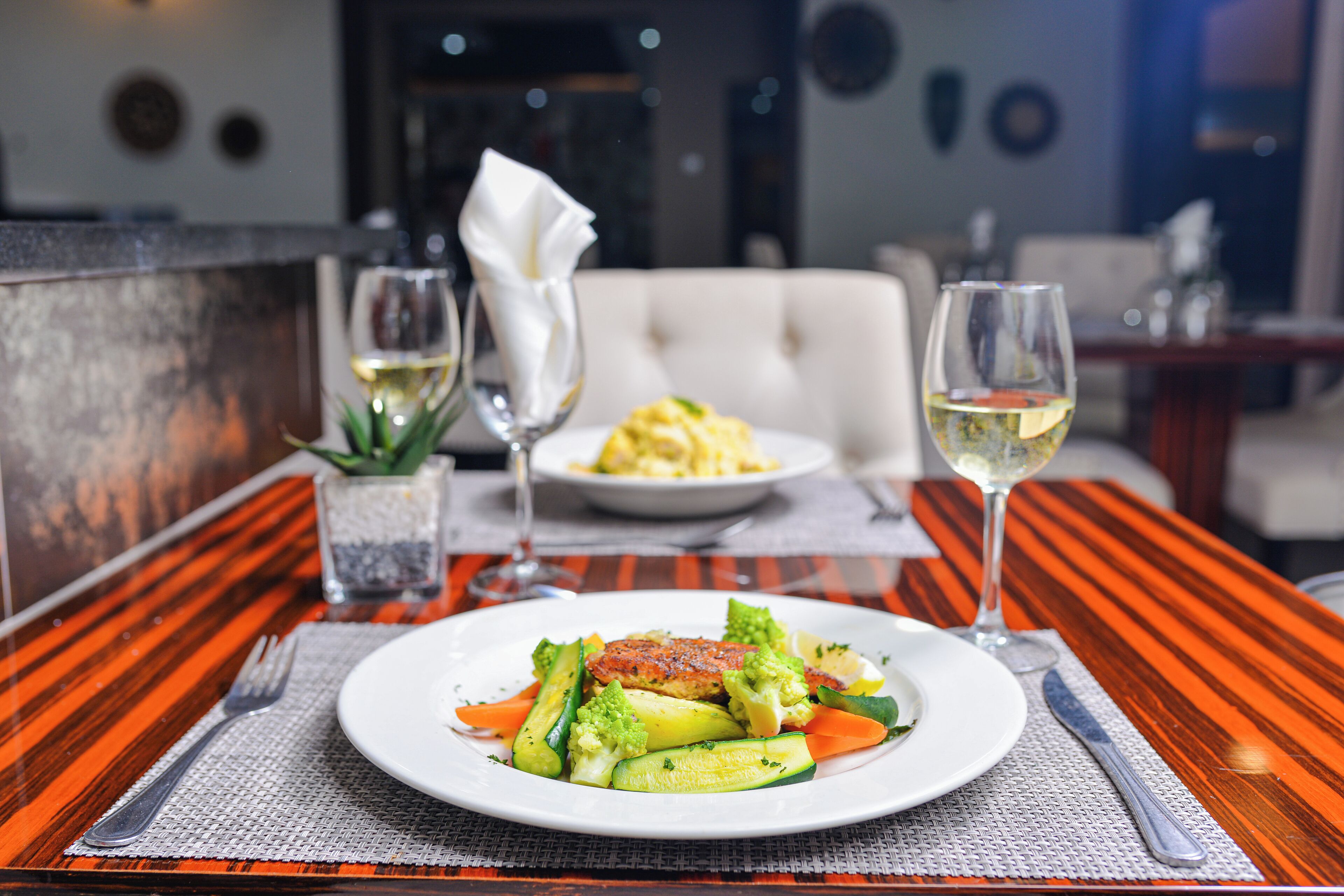 A dining table with a plate of vegetables, a napkin, and a glass of white wine.