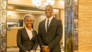 Two people in business attire standing in a lobby with a chandelier and a reception desk in the background.