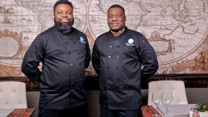 Two chefs in black uniforms standing in front of a world map.