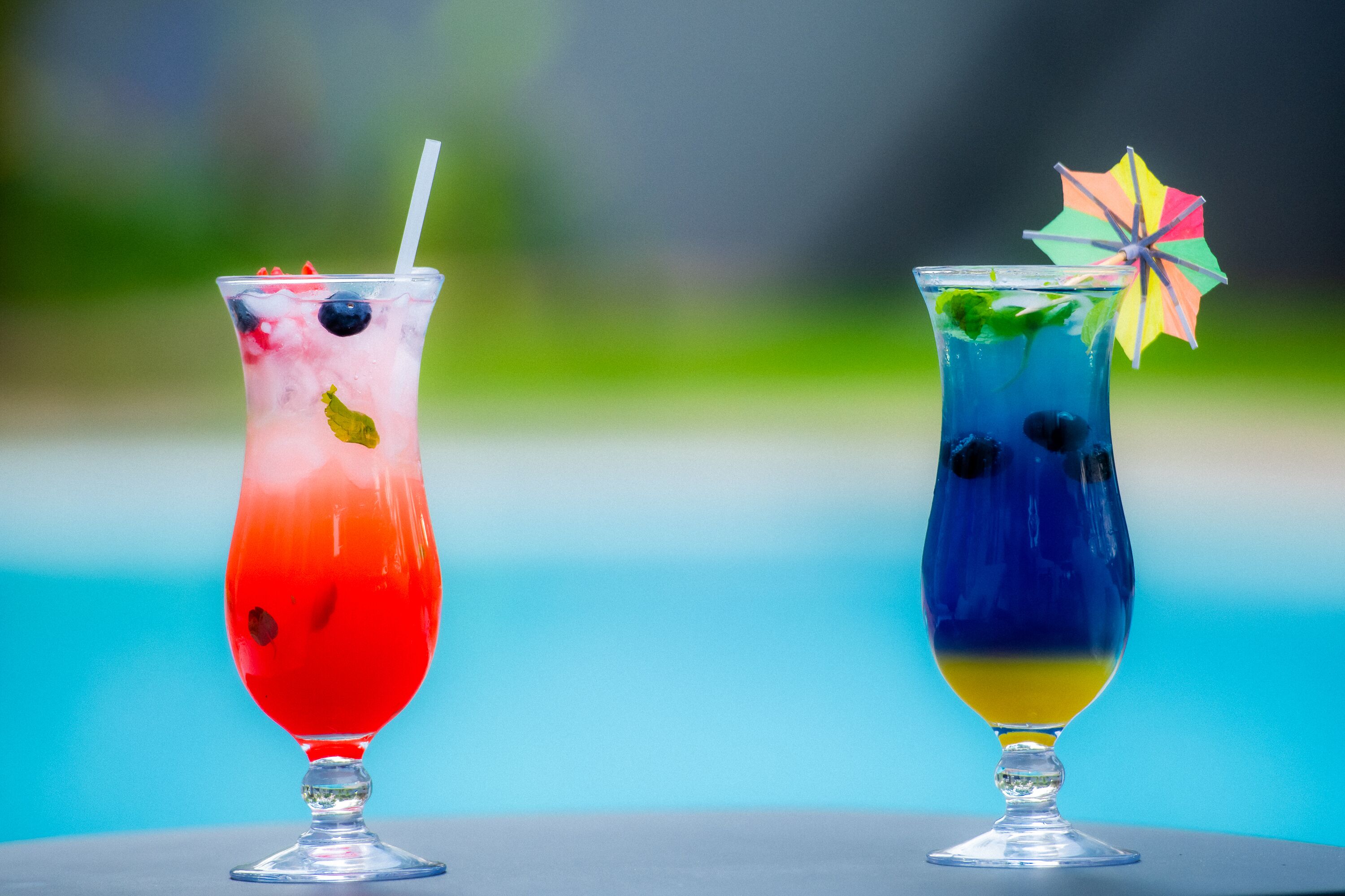 Two cocktails in tall glasses with straws, one with a colorful umbrella, and a blurred outdoor background.