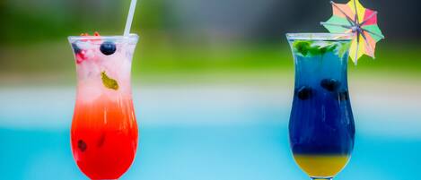 Two cocktails in tall glasses with straws, one with a colorful umbrella, and a blurred outdoor background.