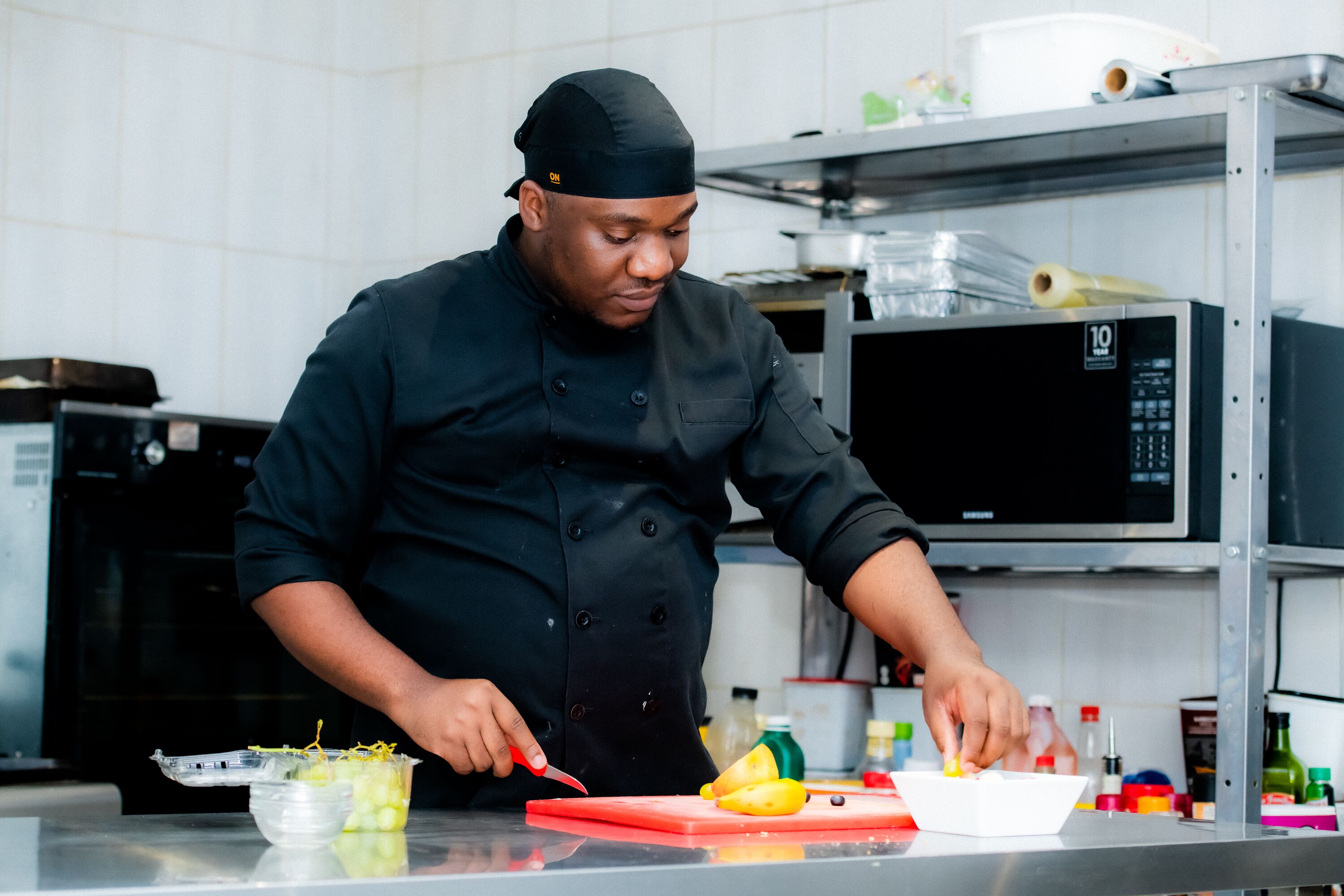 Un chef con uniforme negro corta fruta sobre una tabla de cortar roja en una cocina con electrodomésticos de acero inoxidable y estantes.