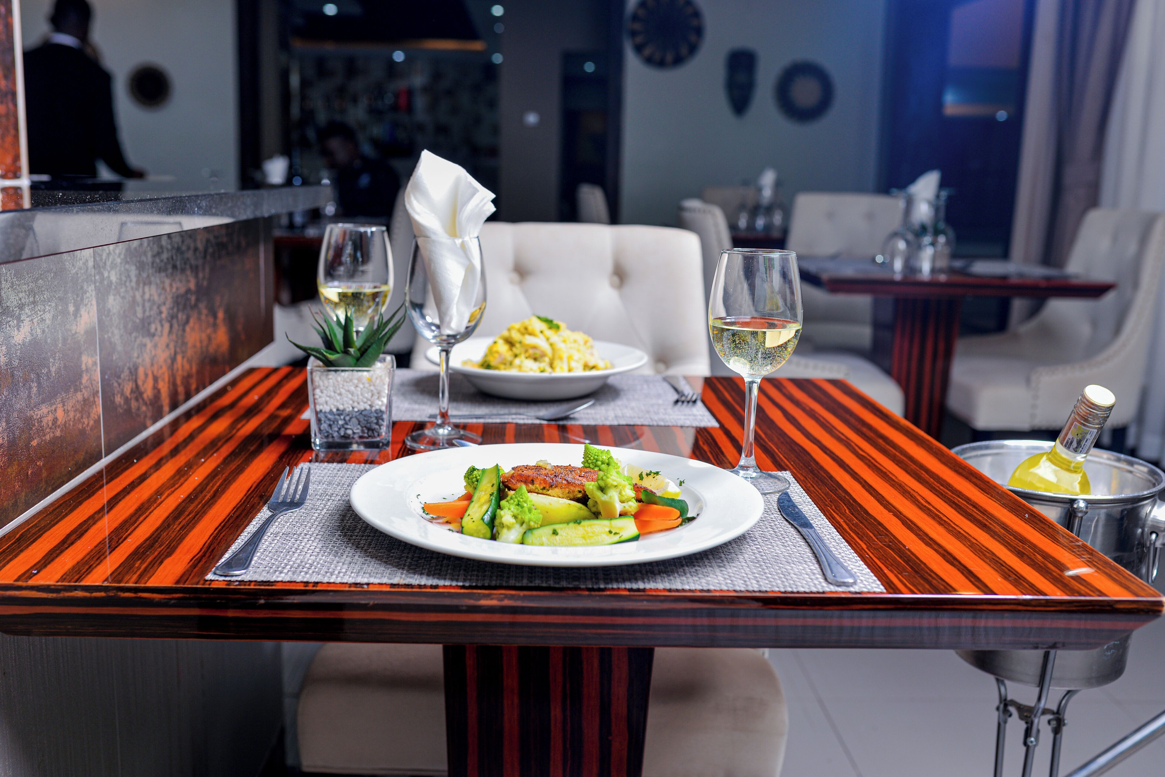 A dining table with a plate of food, two glasses of white wine, and a bottle of champagne.