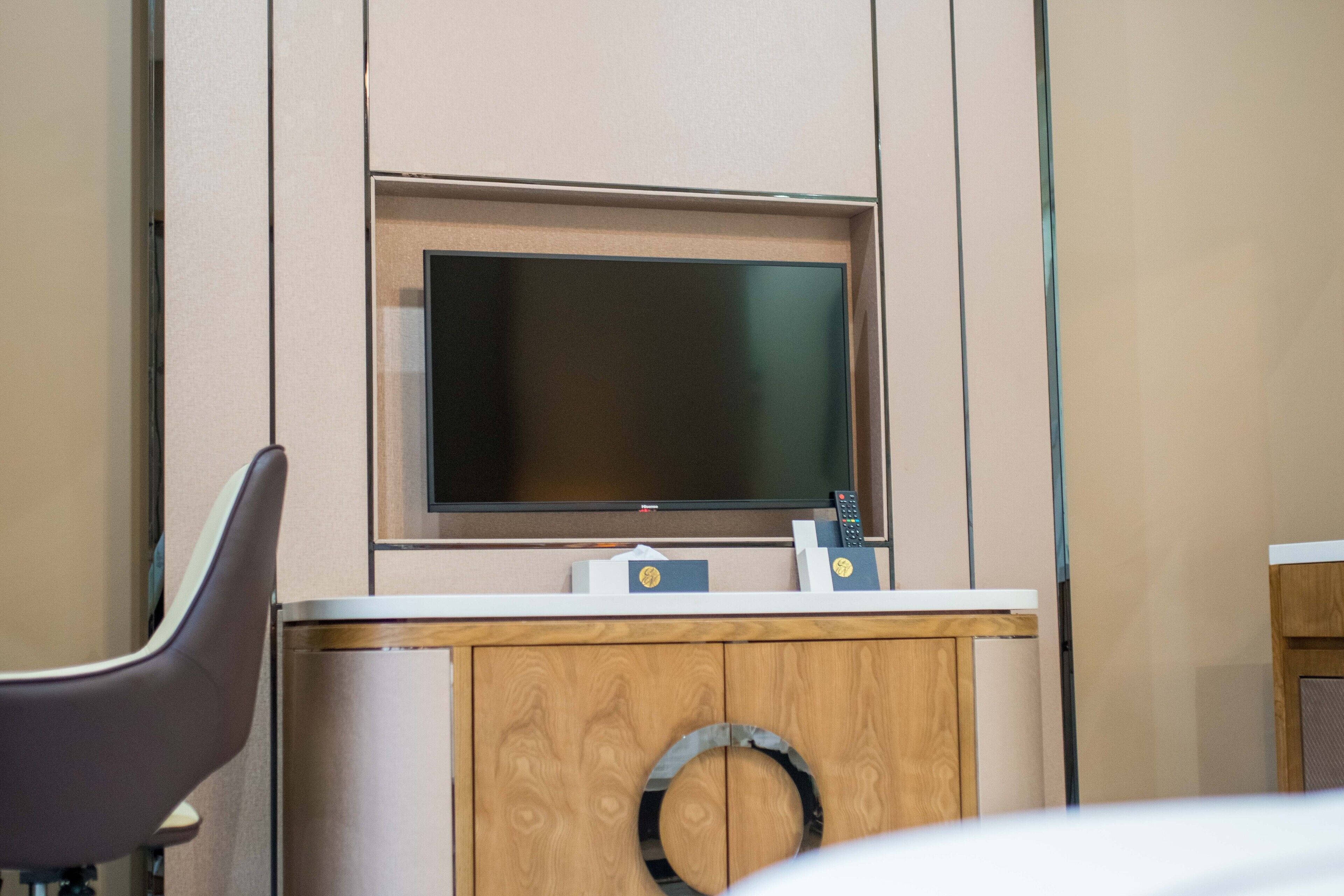 A hotel room with a large flat-screen TV mounted on a wooden cabinet, a desk with a chair, and a small round window.