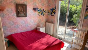 A bedroom with a red bedspread, a radiator, a window with a view of greenery, and a framed picture on the wall.