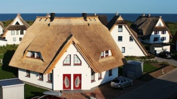 Une maison traditionnelle à toit de chaume, avec une façade blanche et des portes rouges, entourée d’autres maisons similaires et offrant une vue dégagée sur la mer.