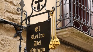 Schild für 'El Retiro Del Obispo' mit einem Kreuzsymbol, einer Glocke und einem hängenden Ornament.