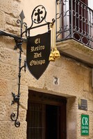 A sign for "El Retiro Del Obispo" with a cross symbol, a bell, and a hanging ornament.
