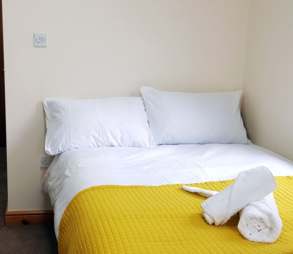 Comfortable bed with a white duvet and two pillows, a yellow quilted bedspread, and a rolled towel.