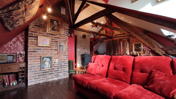 A cozy room with a red velvet sofa, wooden beams, brick walls, and various framed pictures.
