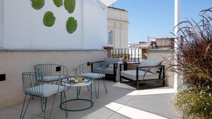 Un área de estar moderna al aire libre con sillas de metal y una mesa, una pared blanca decorada con hojas verdes y un cielo azul despejado.