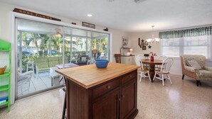 A kitchen with a wooden island, a dining table with chairs, a green shelf, and a sliding glass door leading to a patio with outdoor furniture. - Perfect Island Location! Heated pool and plenty of room for the entire family (Anna Maria)