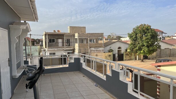 A balcony with a tiled floor, a white railing, and a view of neighboring buildings and a tree. - Captivating 5-bedroom Duplex With 4 Toilets (Brufut)