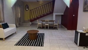 A living room with a sofa, a coffee table, a dining table with chairs, a staircase, and a desk with a lamp. - Captivating 5-bedroom Duplex With 4 Toilets (Brufut)