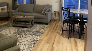 Un salon chaleureux avec un canapé, une table basse et un coin repas avec des chaises. Il y a deux fenêtres avec des rideaux, un plafond en bois et une décoration en forme de boussole fixée au mur.