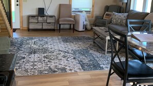 Un vaste salon doté d’un plafond en bois, d’un téléviseur à écran plat fixé au mur, d’un coin salon confortable avec un canapé et des fauteuils, d’une table à manger avec des chaises, et d’un tapis à motifs sur le sol.