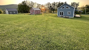 Un paysage rural avec trois maisons, un champ d’herbe et un coucher de soleil en arrière-plan.