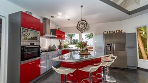A modern kitchen with red and grey cabinetry, a central island with bar stools, a stainless steel refrigerator, and a ceiling light fixture. - Beautiful Carmen Luxury villa (Vitet)