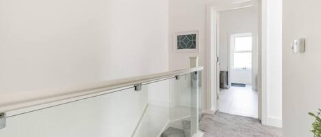Una escalera con barandilla de cristal que lleva a una plataforma, un pequeño cuadro en la pared y una puerta al final del pasillo.