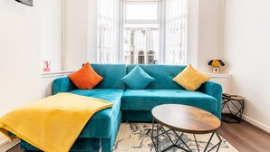 A teal sofa with colorful cushions, a yellow throw blanket, and a round wooden table with metal legs. - Immaculate 4-bed House Near Anfield and Goodison (Bootle)