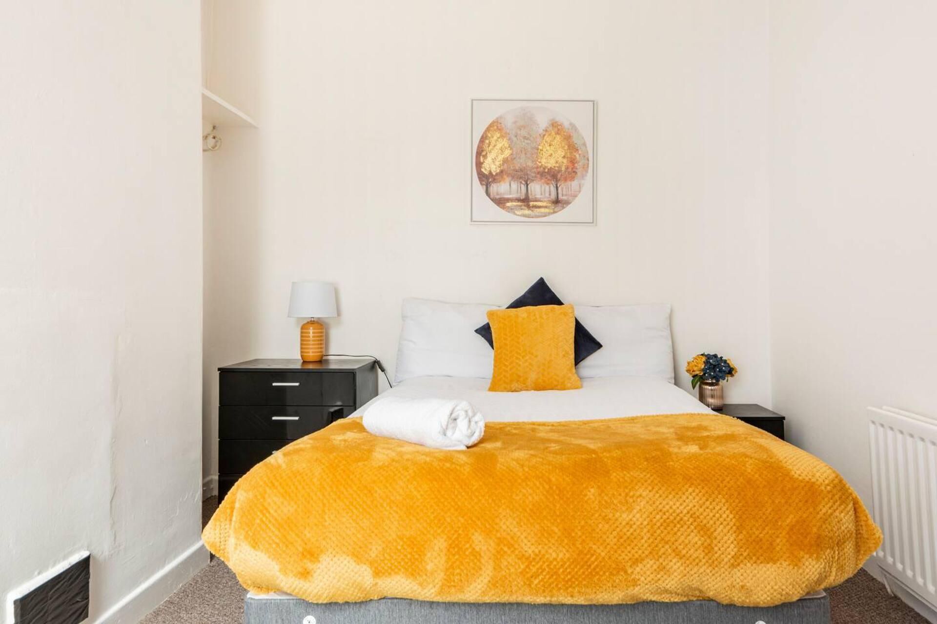 A bedroom with a bed, a nightstand, a lamp, a painting, and a radiator.