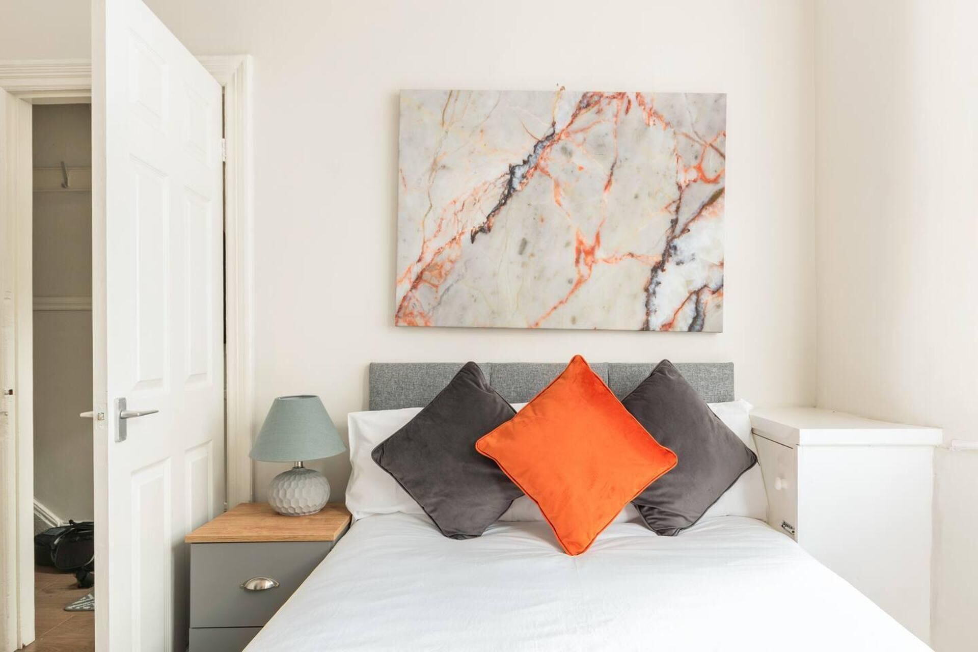A bedroom with a bed, a nightstand with a lamp, a marble artwork on the wall, and a door leading to another room.