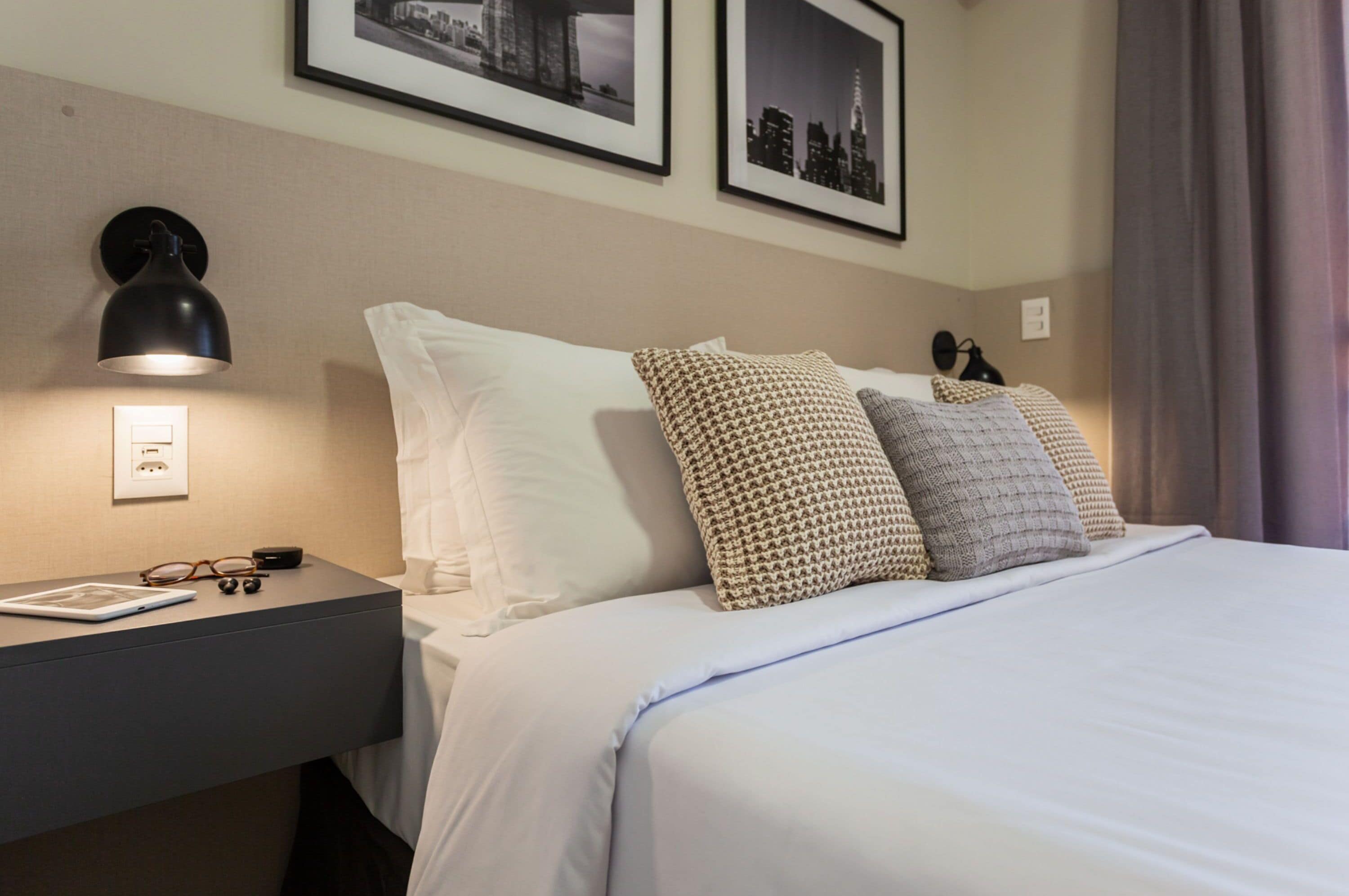 A neatly made bed with white and patterned pillows, a bedside table with glasses and a book, and two framed pictures on the wall.