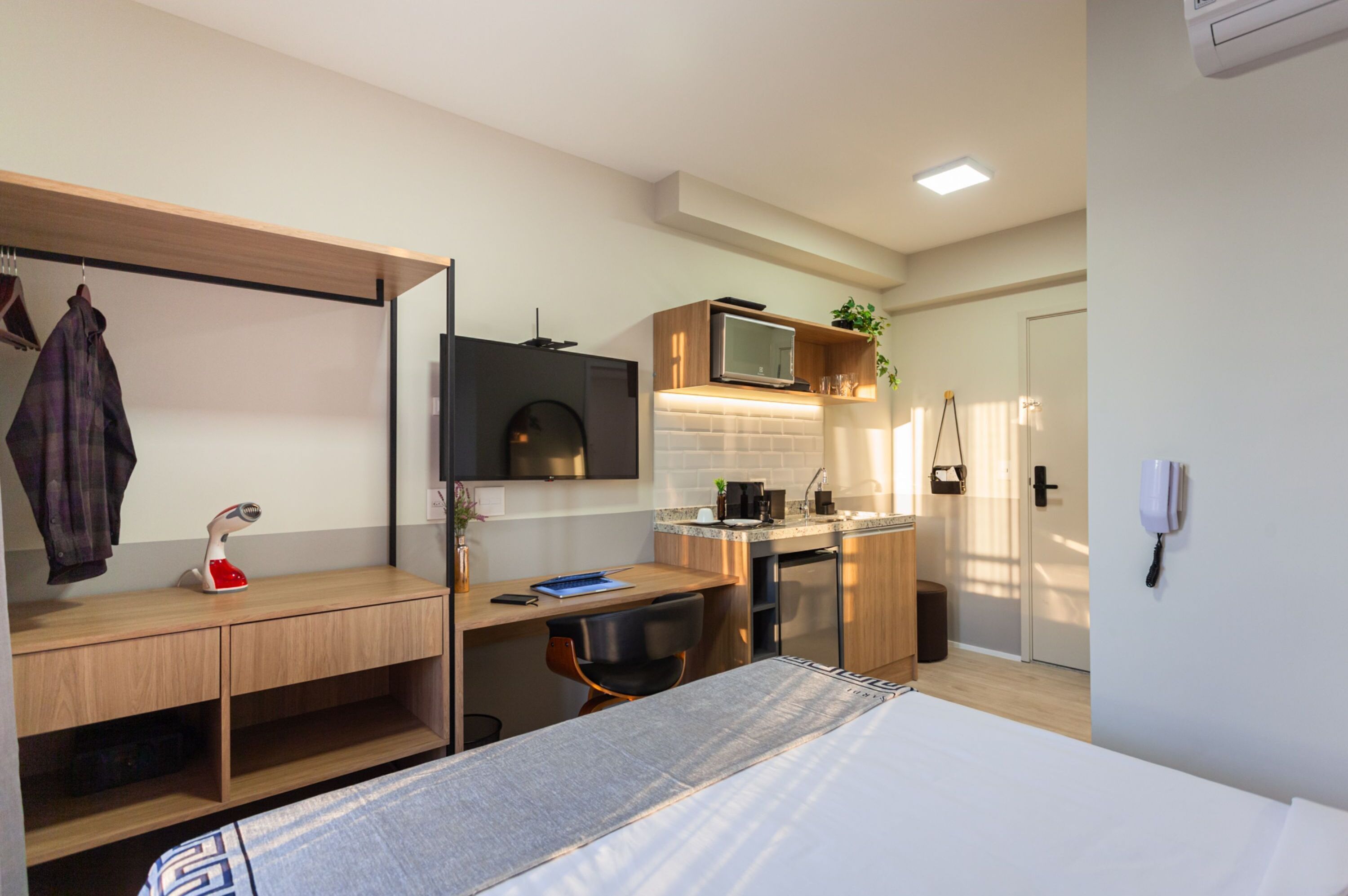 A compact hotel room with a bed, a desk with a chair, a kitchenette, a TV, and a wardrobe.