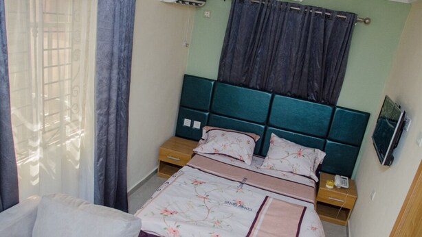 A bedroom with a bed, curtains, a bedside table, and a wall-mounted TV. - Coolio Hotel & Suites (Lagos)