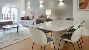 A modern living room with a grey sofa, a round dining table, and four chairs. There are two wall-mounted lamps and a large window with a view of a balcony.