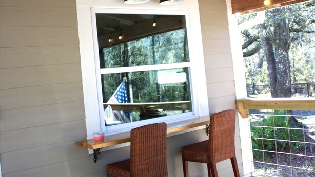 Un porche cubierto con dos sillas de mimbre, una mesa de madera y una ventana con vistas a árboles.