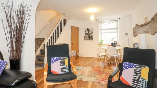 A living room with a staircase, two armchairs with patterned cushions, a dining area with a table and chairs, and a large wooden beam on the wall. - 5 Thomas Street (Caernarfon)
