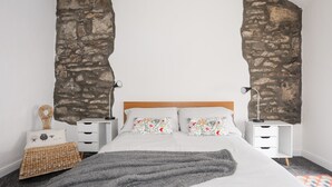 A bedroom with a large bed, two bedside tables, a nightstand, a woven blanket, and a wall with a stone-like texture. - 5 Thomas Street (Caernarfon)