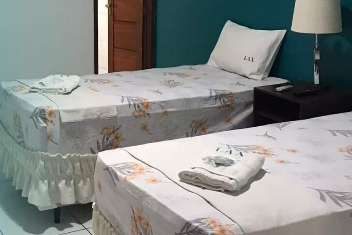 Habitación con dos camas, cada una con una colcha estampada y un almohadón blanco. Se observa una mesita de noche con una lámpara en un lado y una puerta en el otro.