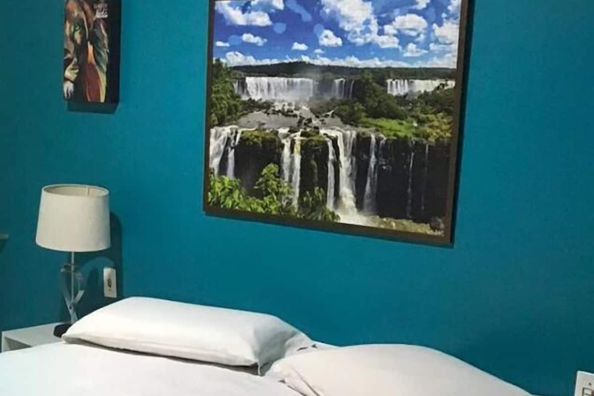 Un dormitorio con cama, almohadas, una lámpara y un cuadro de una cascada en la pared.