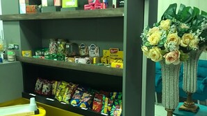 Una botiga on es venen menjar i begudes, amb estantes plenes d'aperitius i refrescos, una taula petita amb una tassa, i gerros decoratius amb flors.