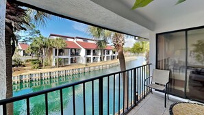 A balcony with a view of a swimming pool, a building with a red roof, and palm trees. - Edgewater Villa #2209 | Lake View, Beach Tram + Resort Amenities (Panama City Beach)