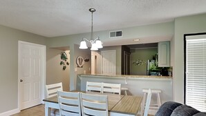 A dining area with a wooden table and white chairs, a kitchen with white cabinets, and a gray couch in the foreground. - Edgewater Villa #2301 | Golf, Beach, 11 Pools! (Panama City Beach)