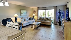 A living room with a sofa, two lamps, a dining table, and a TV. - Edgewater Villa #2301 | Golf, Beach, 11 Pools! (Panama City Beach)