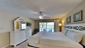Une chambre d’hôtel avec un grand lit, un ventilateur de plafond, une commode blanche, une télévision et un balcon donnant sur la piscine.