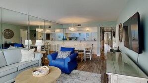 Een moderne woonkamer met een bank, een blauwe fauteuil en een salontafel. In de achtergrond is een keuken te zien met een eettafel en stoelen.